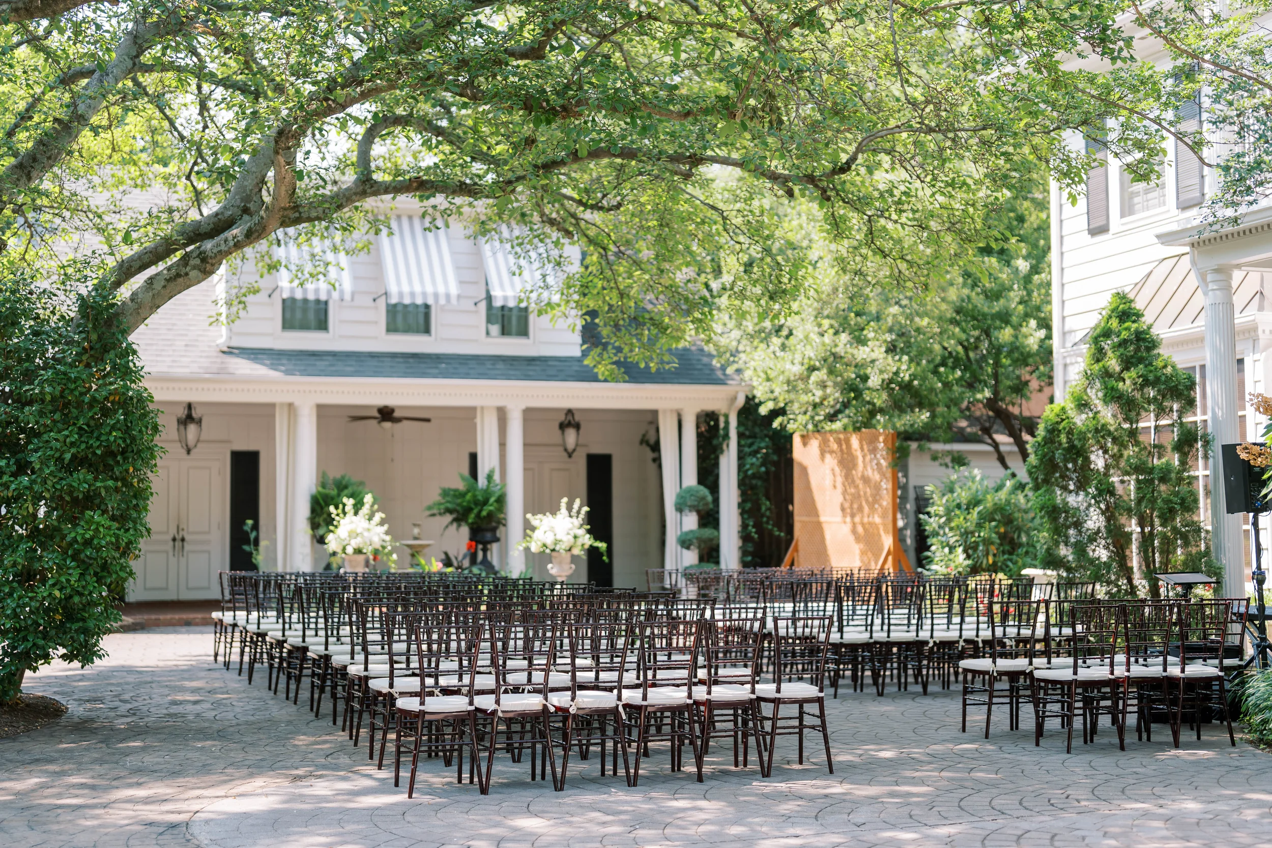 A large white building with a covered porch and columns, surrounded by trees and an outdoor seating area with rows of chairs.